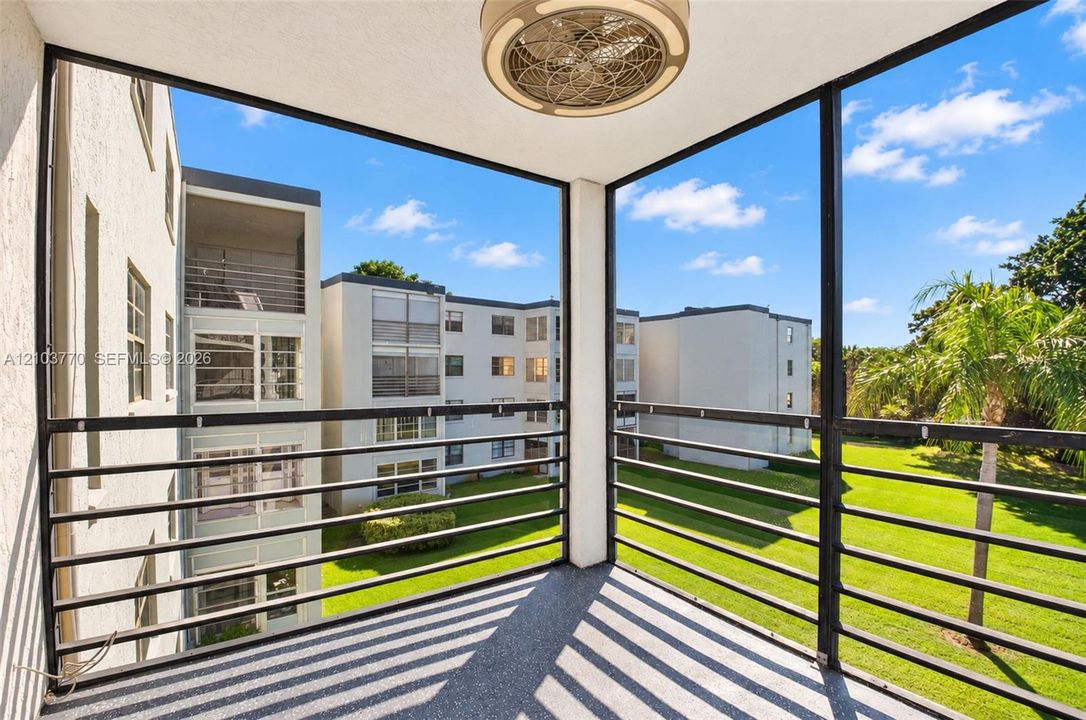 Screened balcony with a mounted ceiling fan, textured flooring, and black metal railings overlooking a landscaped green space