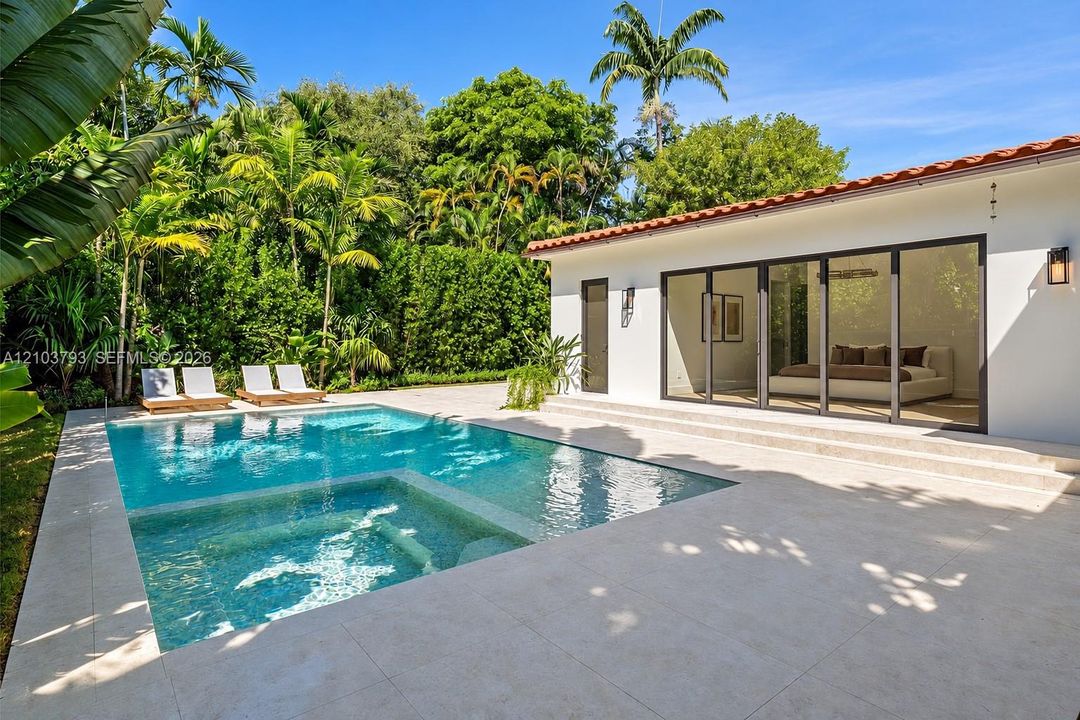 Outdoor swimming pool with integrated spa, expansive light-toned patio, white stucco exterior, multi-panel sliding glass doors, and mature tropical landscaping