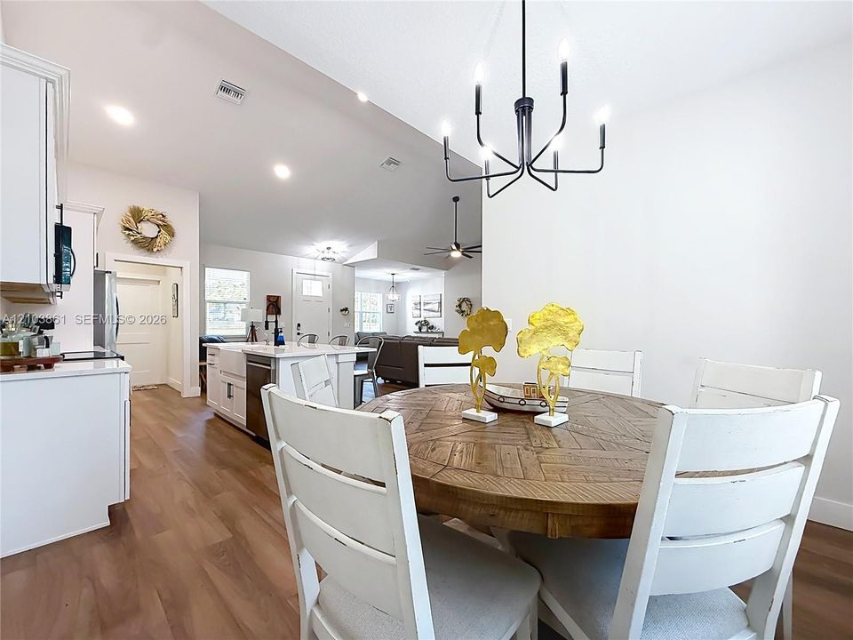 Open-concept kitchen and dining area featuring wood-finish flooring, recessed lighting, matte black multi-arm chandelier, white cabinetry, and a farmhouse-style sink