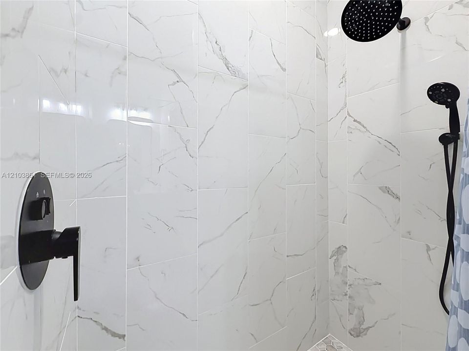 Walk-in shower featuring large-format stone-patterned wall tile, matte black rainfall showerhead, adjustable handheld shower wand, and a matching matte black pressure-balance faucet