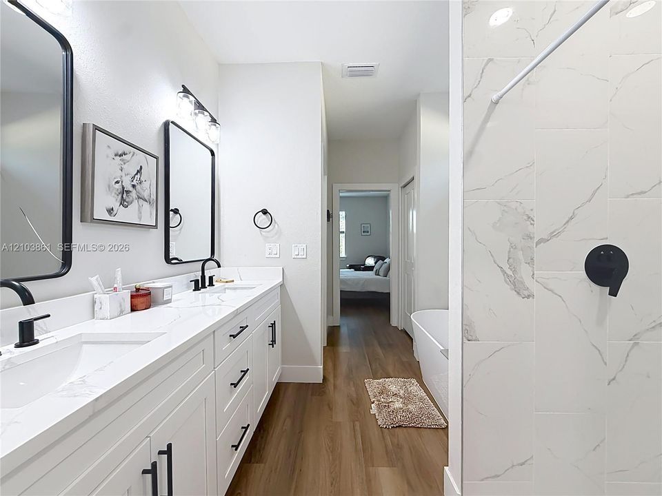 Dual vanity with white cabinetry, stone-finish countertops, matte black fixtures, and framed mirrors, complemented by wood-finish flooring and a tiled shower surround