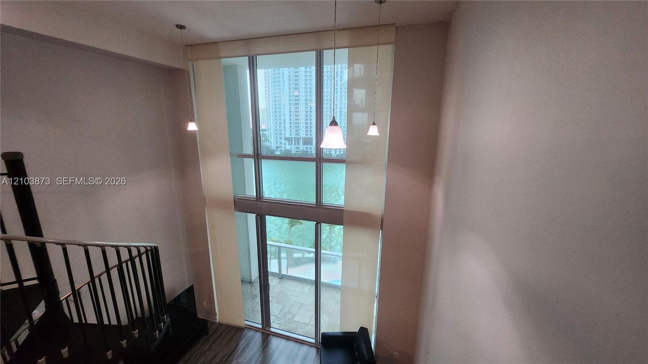 Double-height floor-to-ceiling windows, suspended pendant lighting, dark metal spiral staircase with railing, and wood-finish flooring