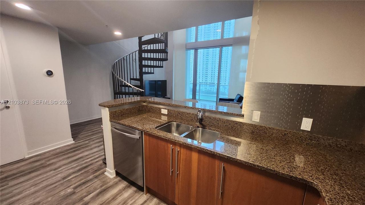 Kitchen featuring stone countertops, stainless steel dual-basin sink, wood-finish cabinetry, and stainless steel dishwasher, complemented by a metal spiral staircase and floor-to-ceiling windows
