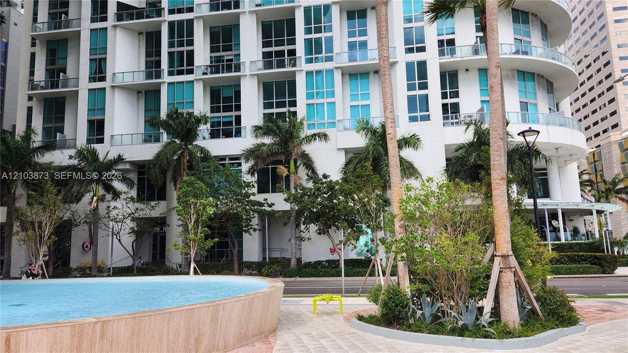 High-rise exterior featuring curved architectural design, floor-to-ceiling glass windows, private balconies, a circular water feature, and landscaped grounds with mature palm trees