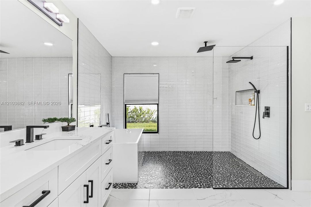 White vanity with stone countertop, freestanding bathtub, walk-in shower with river stone flooring, glass enclosure, and matte black fixtures