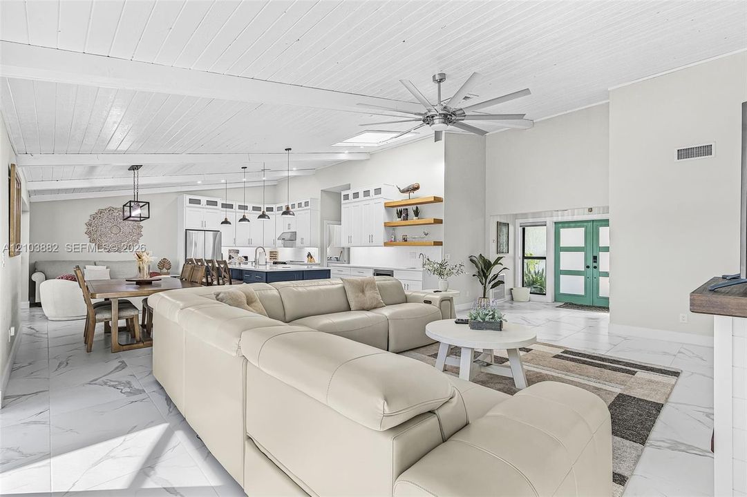 Open-concept interior featuring vaulted ceilings with exposed beams, large-format tile flooring, kitchen with white cabinetry and a contrasting center island, skylight, and multi-pane entry door