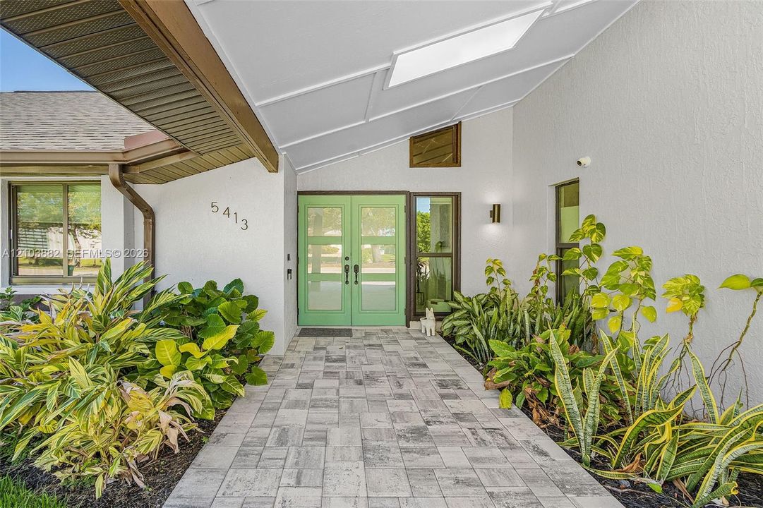 Covered entryway featuring a double-door entry, geometric transom window, stone-finish paver walkway, and exterior wall-mounted lighting