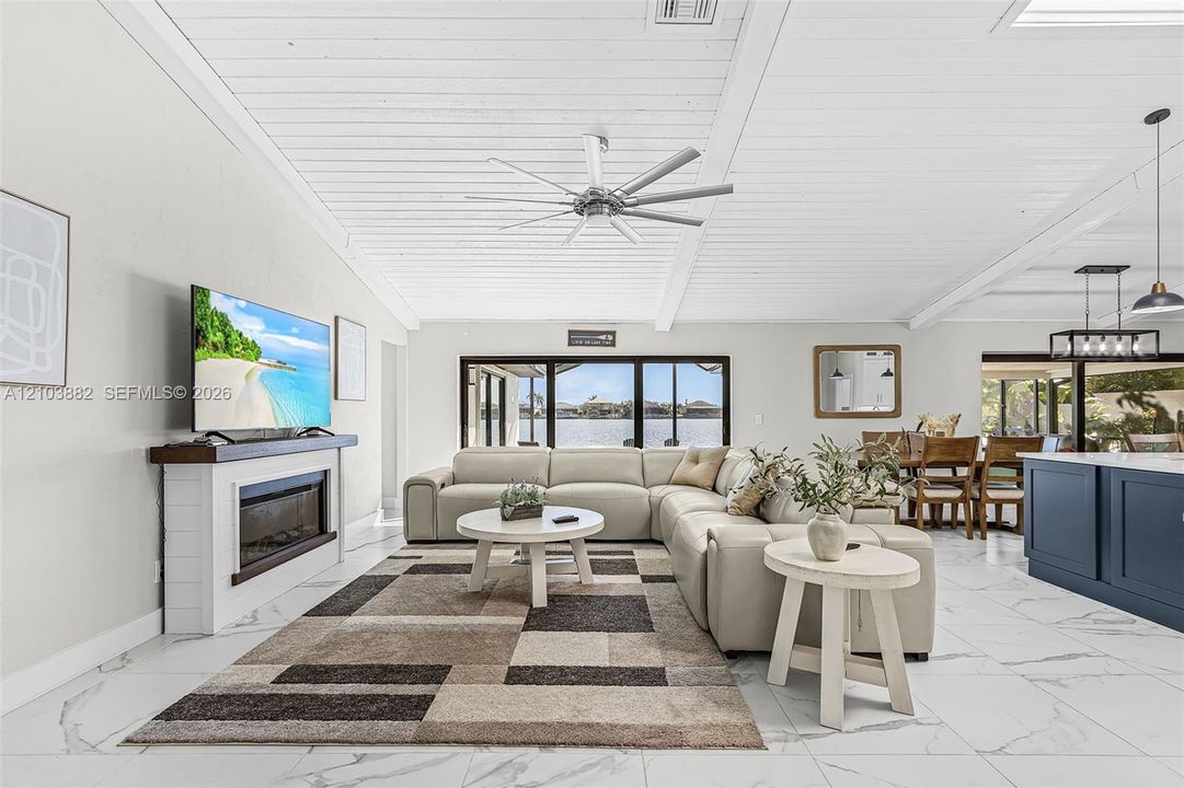 Vaulted ceilings with exposed beams, large format stone-finish flooring, floor-to-ceiling sliding glass doors, built-in fireplace with a timber mantel, and modern multi-blade ceiling fan