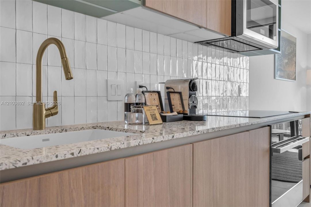 Kitchen featuring speckled composite countertops, wood-finish cabinetry, a white rectangular undermount sink, a brass-tone gooseneck faucet, and a white vertical tiled backsplash
