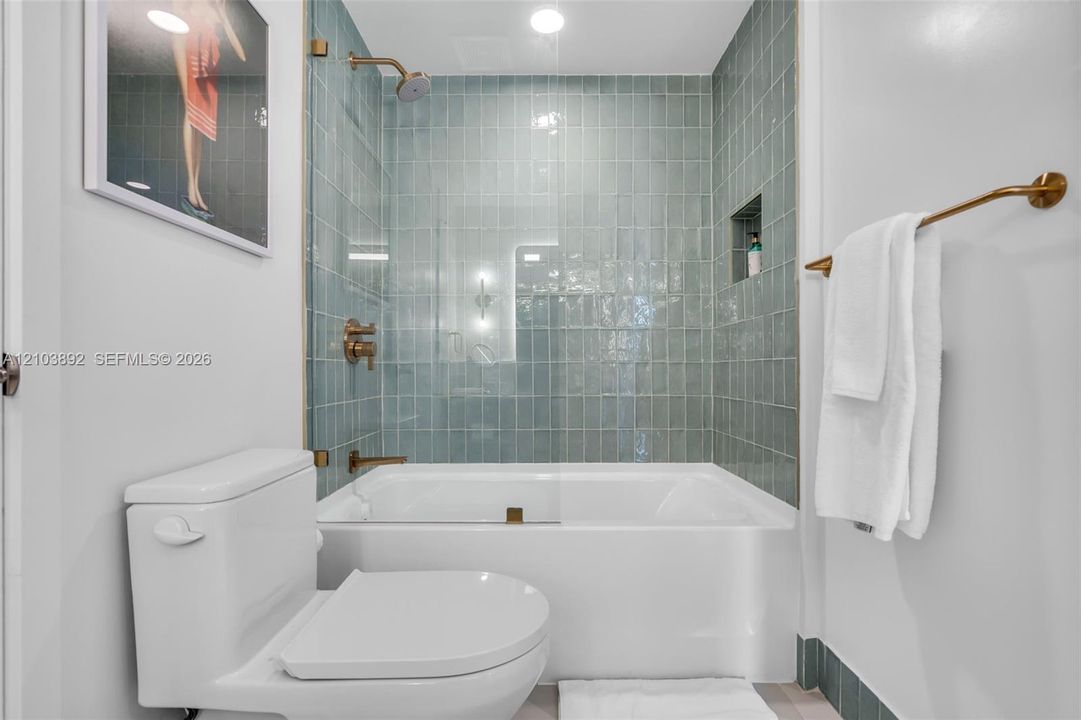 Bathtub and shower combination featuring a glass panel, blue-toned subway wall tile, brushed gold-tone fixtures, and a recessed tiled niche