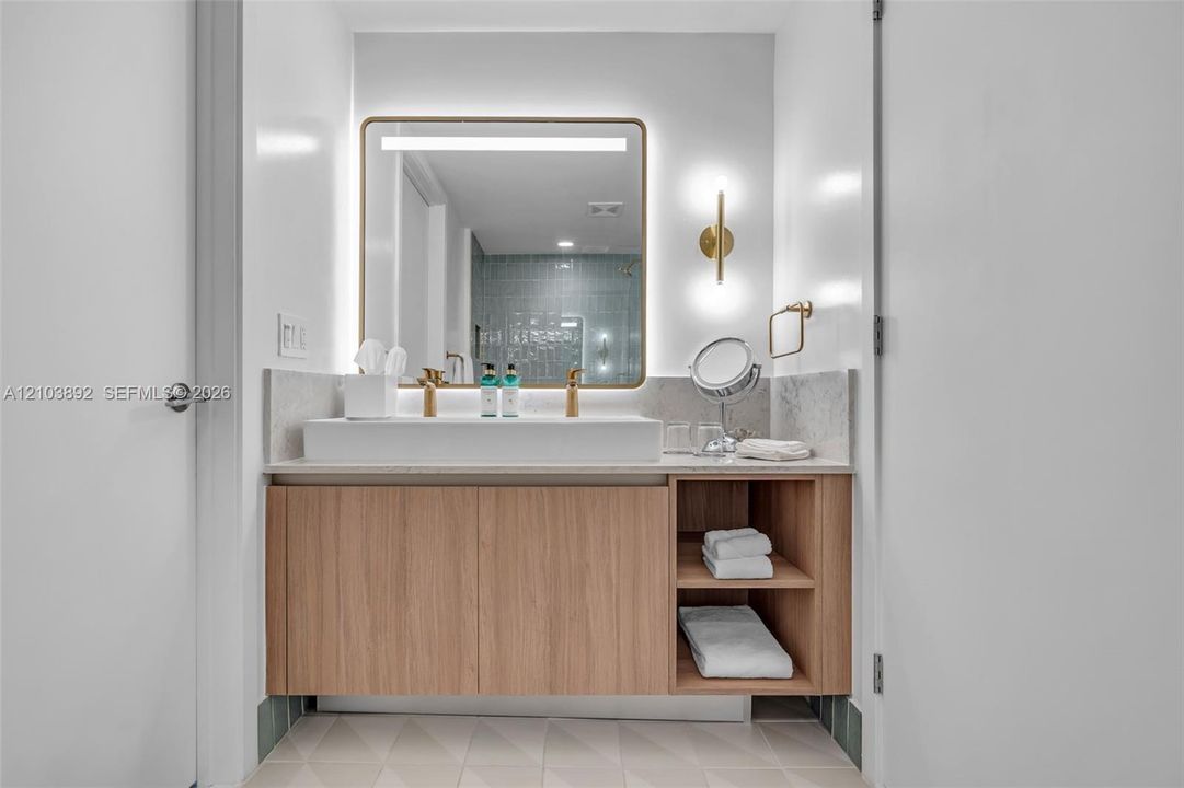 Floating vanity featuring wood-finish cabinetry with open shelving, rectangular vessel sink, stone countertop, integrated LED-backlit mirror, and metallic wall-mounted light fixtures