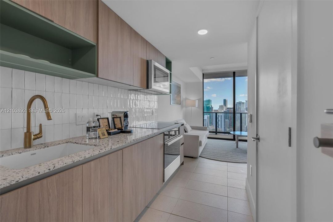 Kitchen featuring wood-finish cabinetry, stone-flecked countertop, white textured tile backsplash, brass-toned faucet, and built-in stainless steel appliances