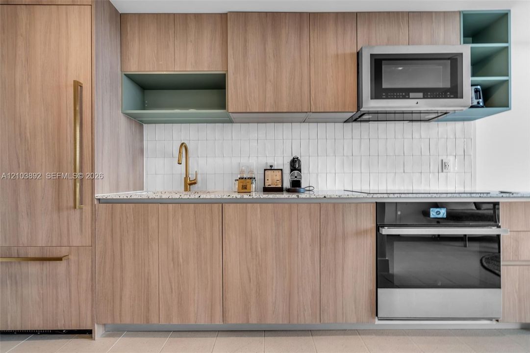 Modern kitchen featuring flat-panel wood-finish cabinetry, a white vertically-tiled backsplash, brass-toned faucet, integrated stainless steel appliances, and a speckled stone countertop