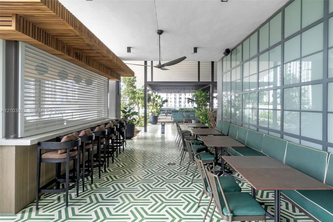 Outdoor covered lounge area featuring geometric patterned tile flooring, a built-in bar with wood-slat soffit detail, and industrial-style metal-framed glass wall partitions