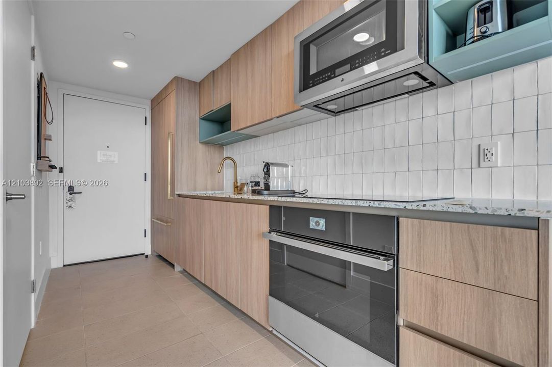 Kitchen featuring wood-finish cabinetry, speckled stone countertop, white square tile backsplash, integrated stainless steel appliances, and a brass-tone gooseneck faucet
