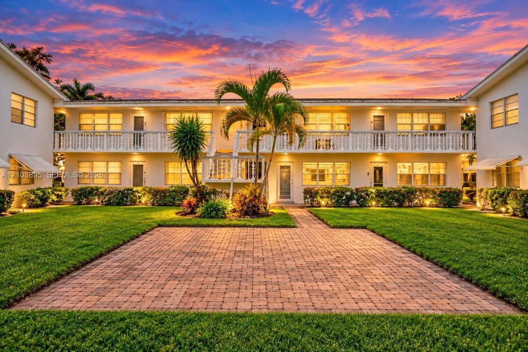 Two-story building exterior featuring a paver stone patio, manicured lawn, second-story balcony with decorative railing, and mature landscaping