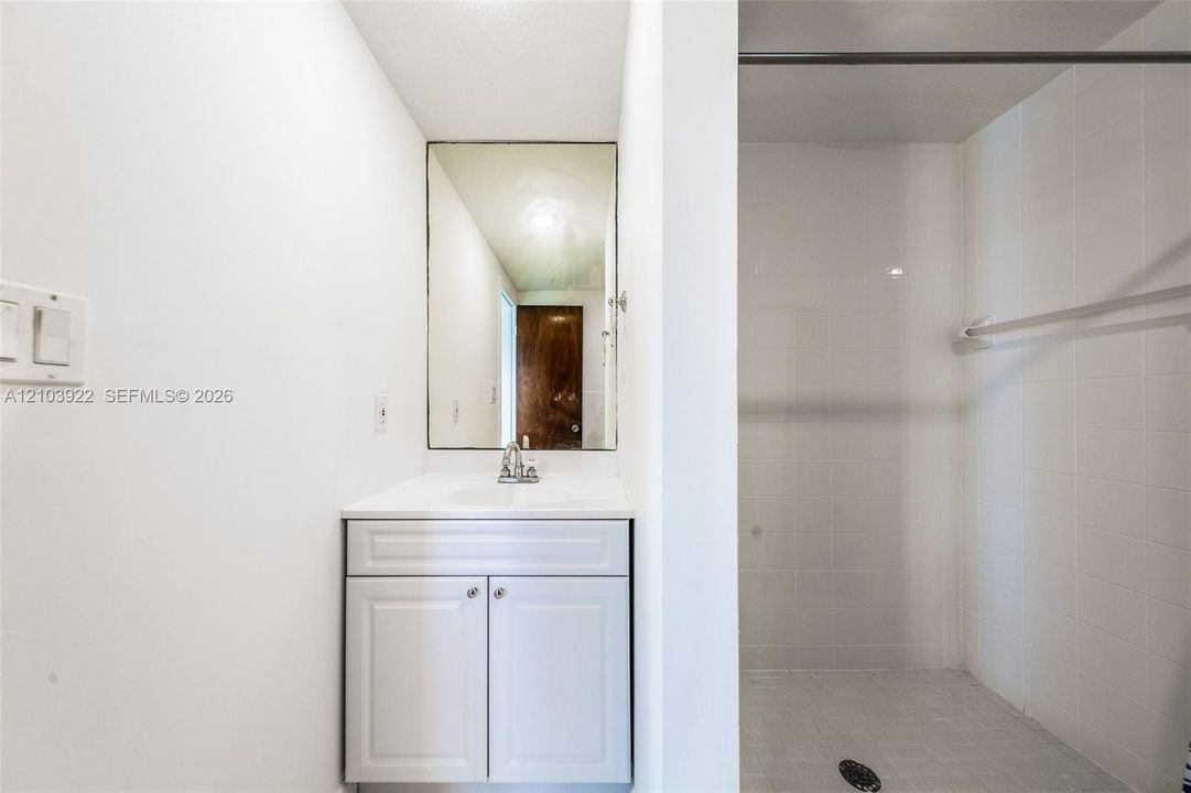 White vanity with a rectangular sink, wall-mounted mirror, and walk-in shower featuring white tiled walls and floor