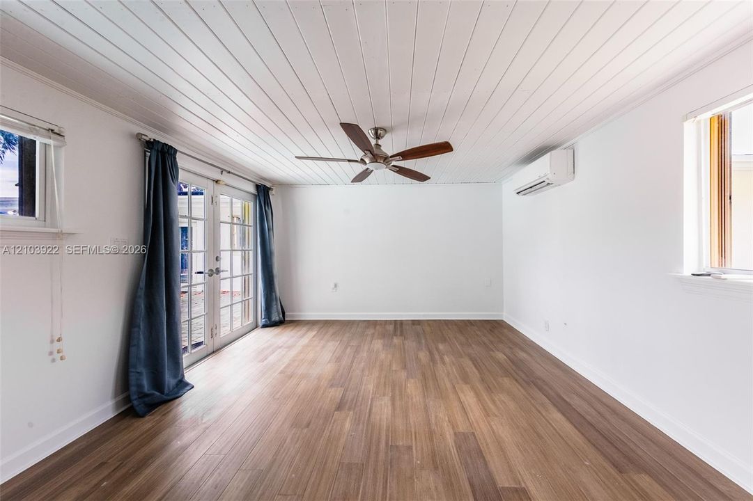 Room featuring wood-finish flooring, white plank-style ceiling, ceiling fan, wall-mounted ductless air conditioning unit, and glass-paneled double doors