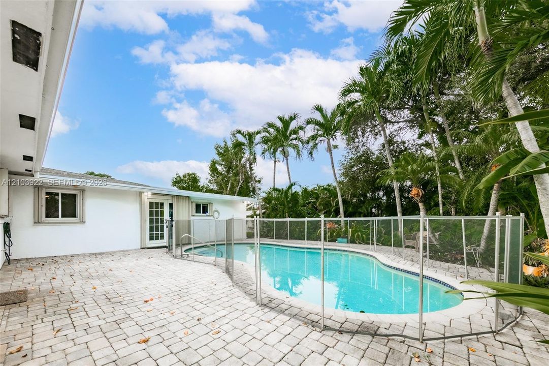 White exterior with an attached patio featuring paver flooring, a fenced in-ground swimming pool, and mature palm trees