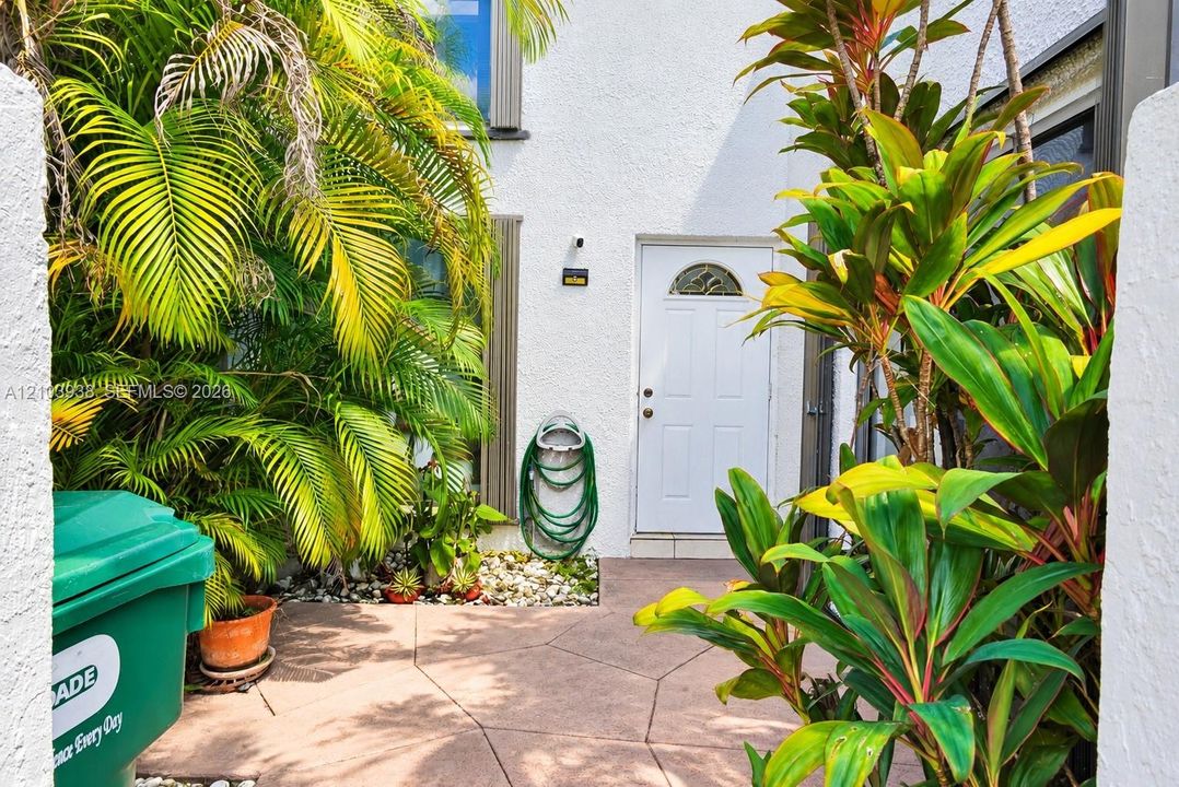 Stucco exterior featuring an arched-top entry door, stone-finish paving, and mature tropical landscaping
