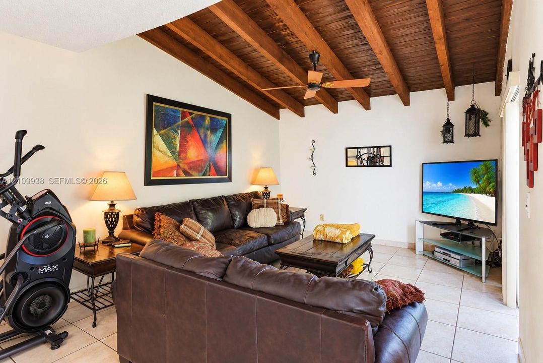 Vaulted ceiling featuring exposed wood beams, neutral floor tiling, and a ceiling fan with integrated lighting
