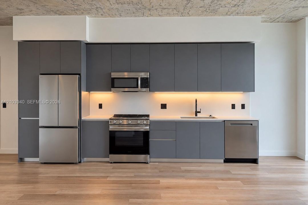 Kitchen featuring matte grey flat-panel cabinetry, light-toned countertops, stainless steel appliances, under-cabinet lighting, and wood-finish flooring