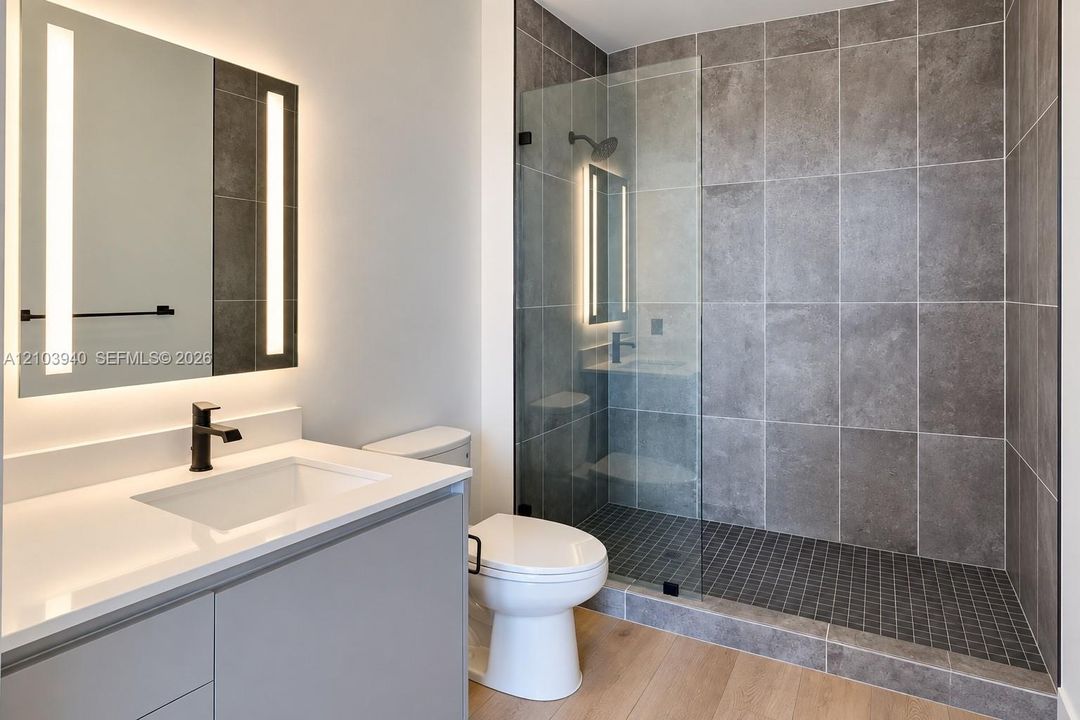 Bathroom featuring a vanity with a white countertop, wall-mounted mirror with integrated lighting, glass-enclosed walk-in shower with grey wall tile, wood-finish flooring, and matte black hardware