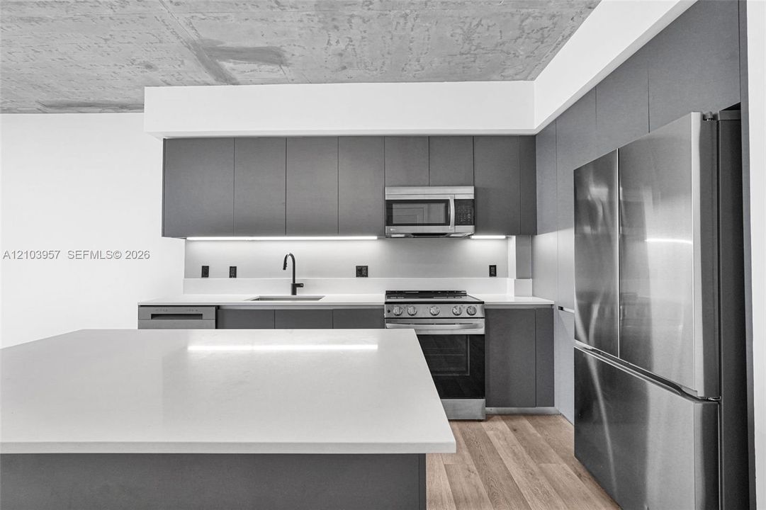 Kitchen featuring flat-panel cabinetry, stainless steel appliances, quartz countertops, wood-finish flooring, and under-cabinet lighting
