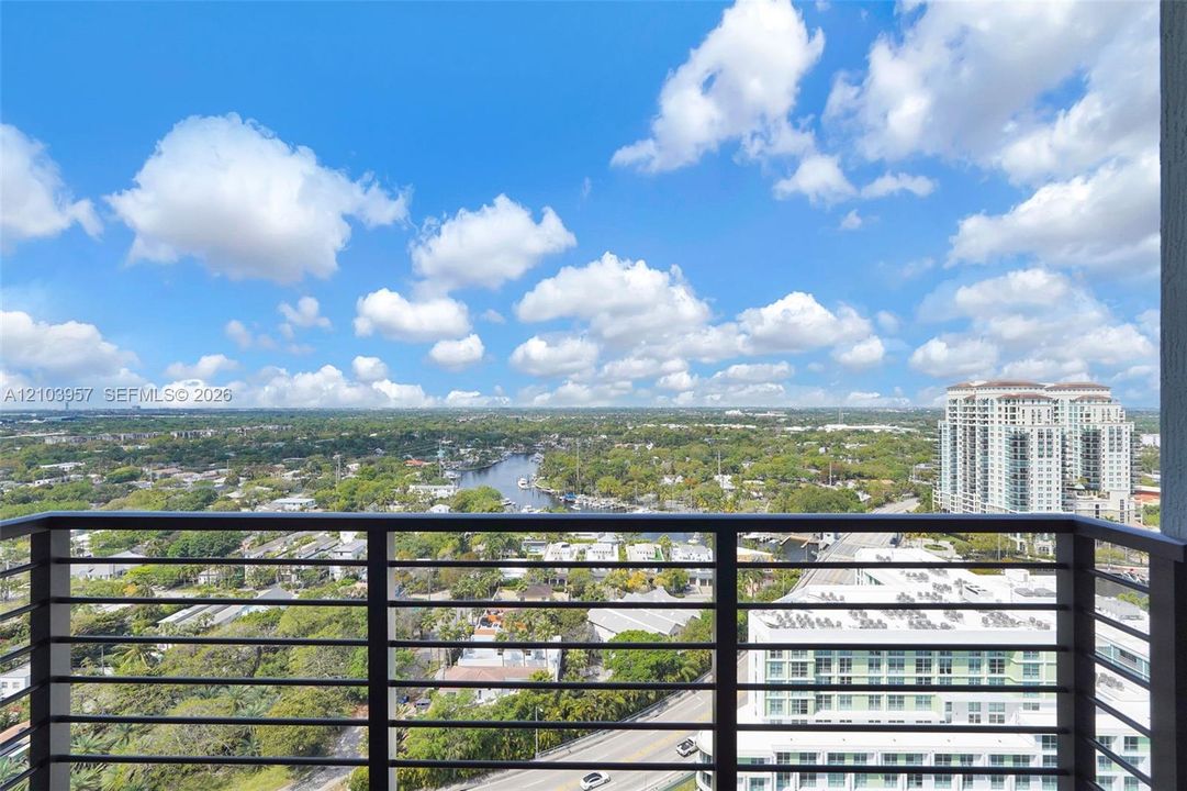 High-rise balcony with horizontal metal railings, overlooking a waterway, urban landscape, and distant buildings
