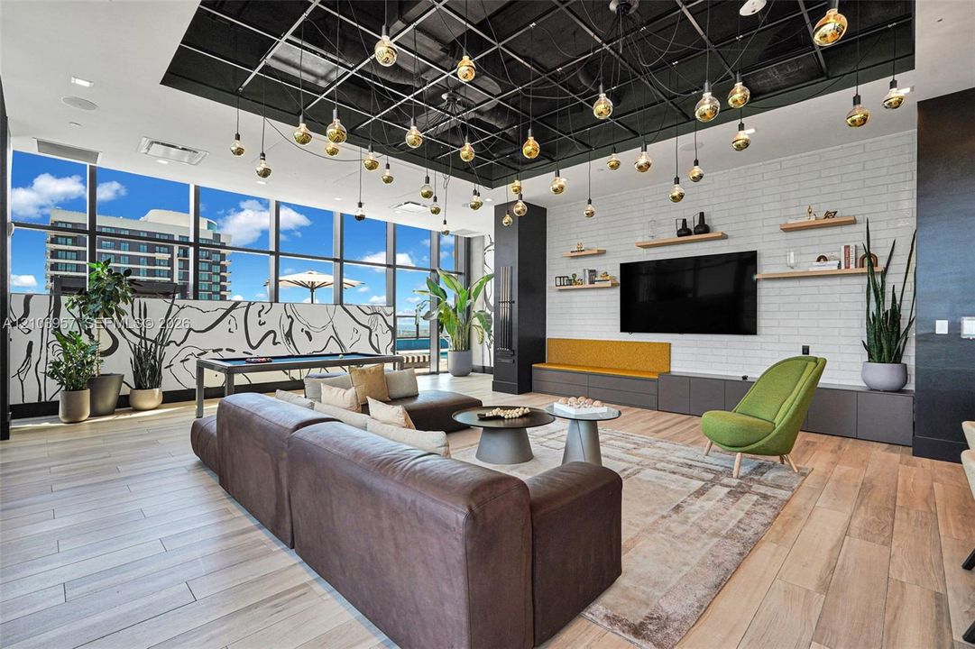 Expansive living area with large windows, wood-finish flooring, white brick-patterned accent wall, built-in shelving, and a unique grid-patterned ceiling with multiple pendant lights