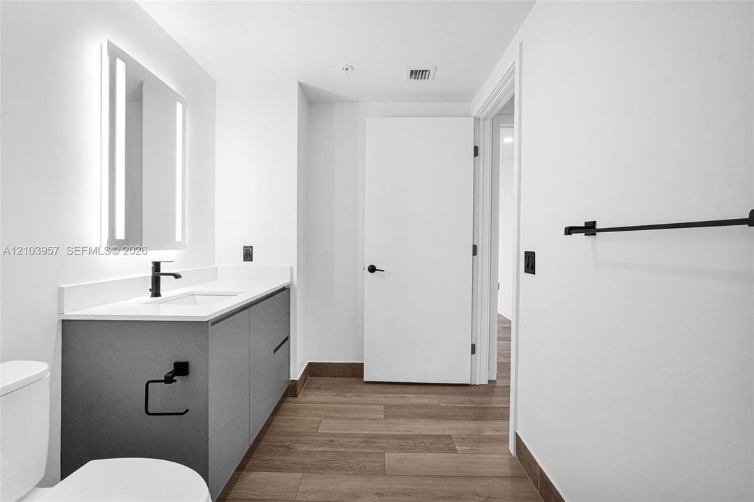 Contemporary bathroom featuring a floating vanity with a white countertop and integrated sink, an illuminated mirror, matte black fixtures, and wood-finish flooring