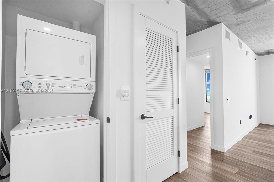 Stacked laundry appliances, louvered interior door, wood-finish flooring, exposed concrete ceiling, and white wall finishes