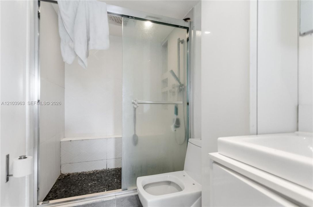 Shower enclosure with glass sliding doors, built-in tiled bench, stone-textured flooring, wall-mounted shower fixture, and white vanity with integrated basin