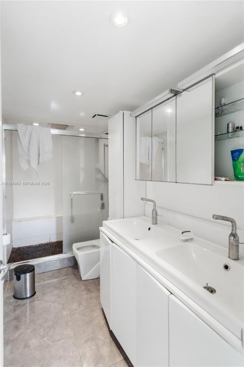 Dual-sink vanity with integrated basins and satin-finish faucets, mirrored medicine cabinetry, and a walk-in glass-enclosed shower with stone flooring