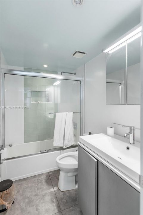 Bathroom featuring a vanity with an integrated sink, metal-finish faucet, medicine cabinet, stone-finish tile flooring, and a tub-shower combination with a sliding glass door and tiled surround