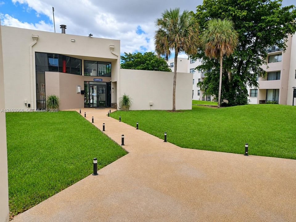 Stucco facade with floor-to-ceiling glass entryway, aggregate concrete walkway, bollard lighting, and landscaped lawn with mature palms