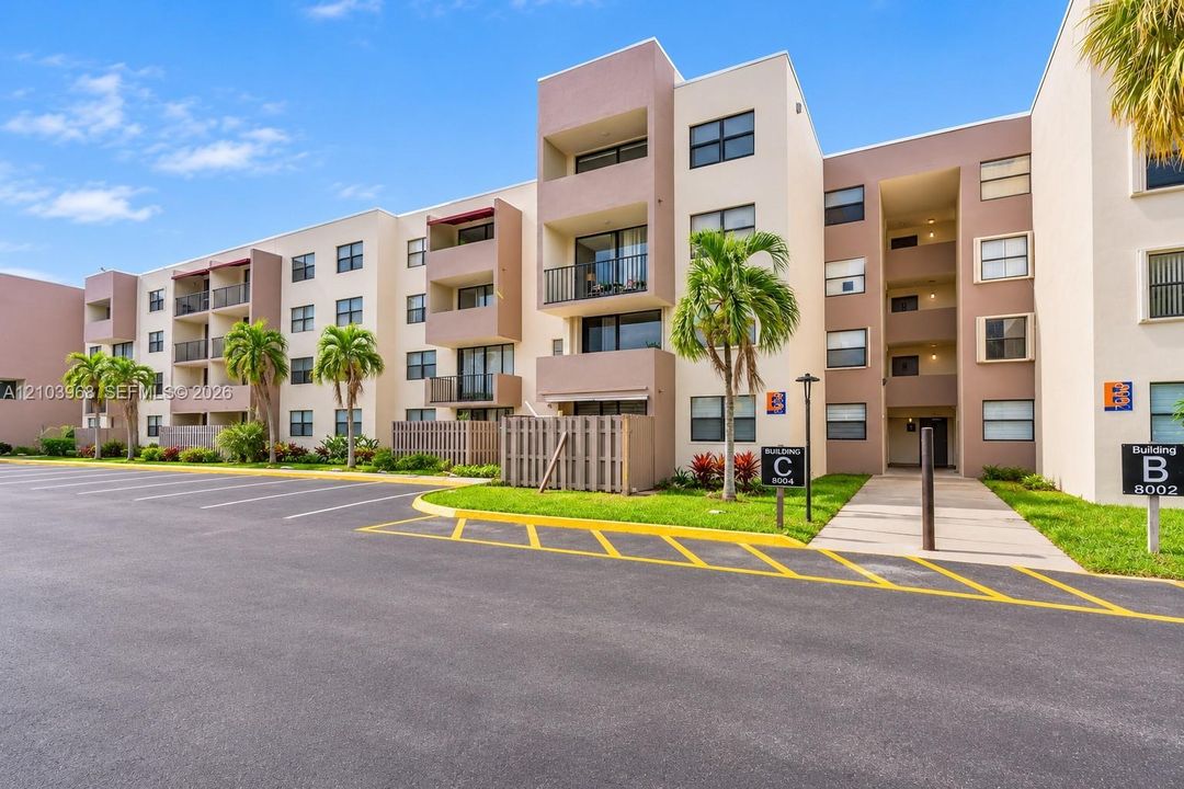 Multi-story residential building exterior with neutral-toned stucco facade, private balconies, paved parking area, and manicured landscaping with mature palms