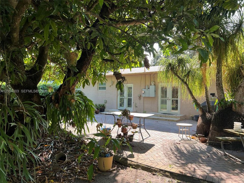 Rear exterior featuring a herringbone-patterned brick paver patio, two sets of white-framed glass French doors with shutters, and mature shade trees