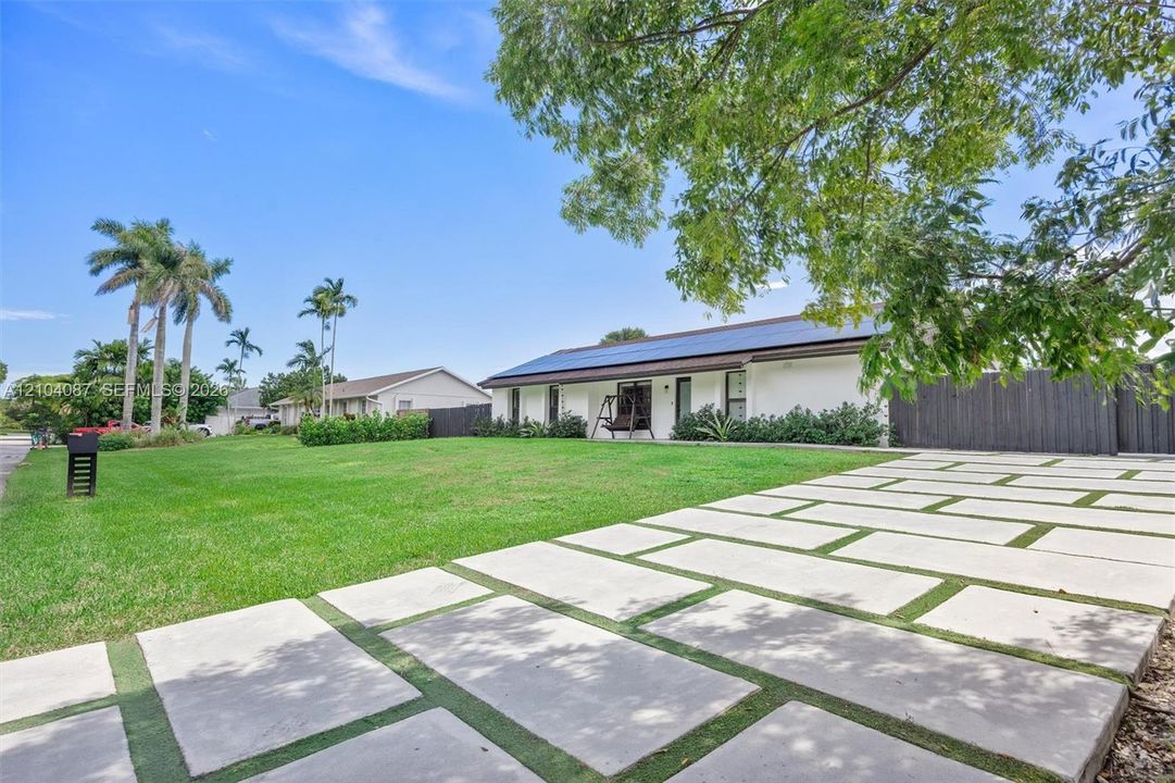 Modern exterior with solar panels, a concrete paver driveway, expansive green lawn, and mature trees
