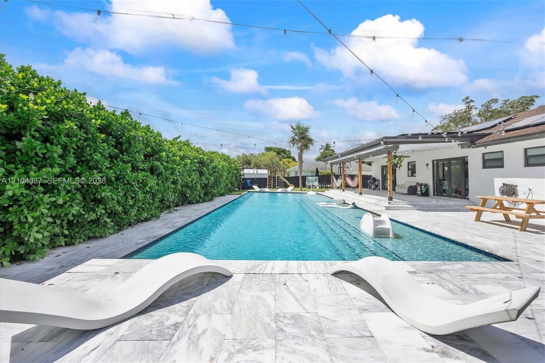 Expansive outdoor living area featuring a rectangular swimming pool, large patio with light-toned pavers, and a covered patio structure