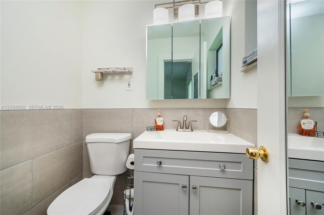 Grey vanity cabinet with rectangular integrated sink, neutral wall tile wainscoting, triple-light fixture, and three-panel mirrored medicine cabinet