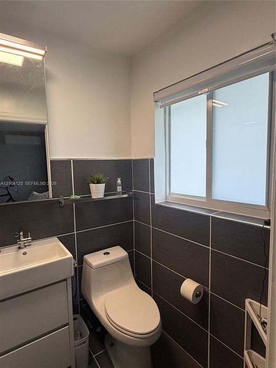 Bathroom featuring floor-to-ceiling charcoal-toned wall tiling, a white rectangular sink with a chrome-finished faucet, a high-profile one-piece toilet, and a sliding window with a integrated roller blind