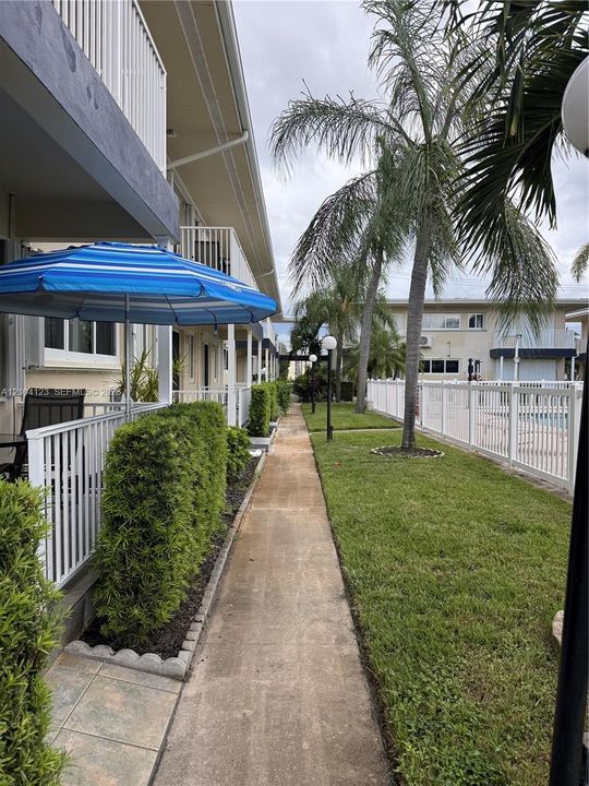 Concrete pathway flanked by manicured landscaping, mature palm trees, white perimeter fencing, and exterior balconies