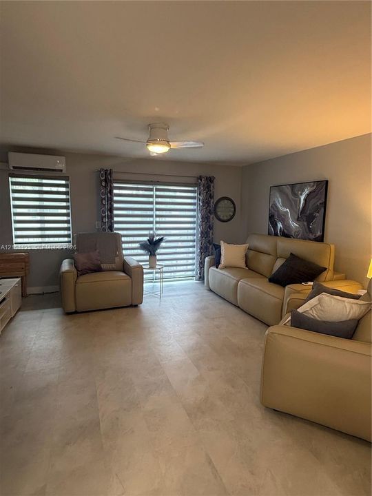 Living area featuring neutral-tone flooring, ceiling fan with integrated lighting, wall-mounted ductless climate control system, and sliding glass doors with dual-layer window treatments