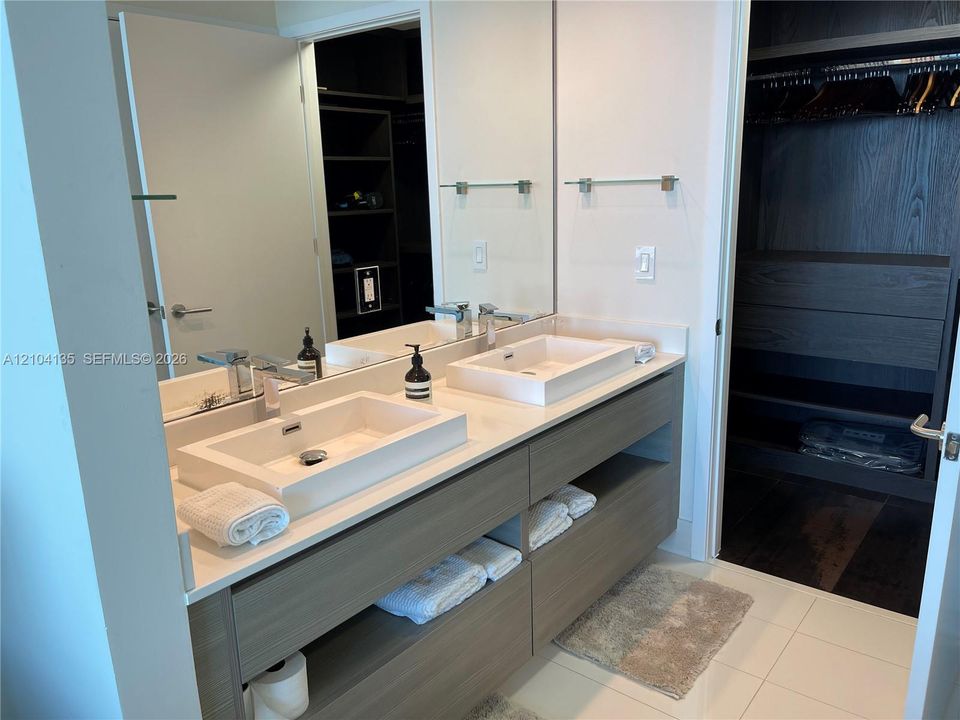 Floating dual vanity with rectangular vessel sinks, quartz countertops, and built-in shelving, complemented by a large wall-mounted mirror and walk-in closet with custom shelving