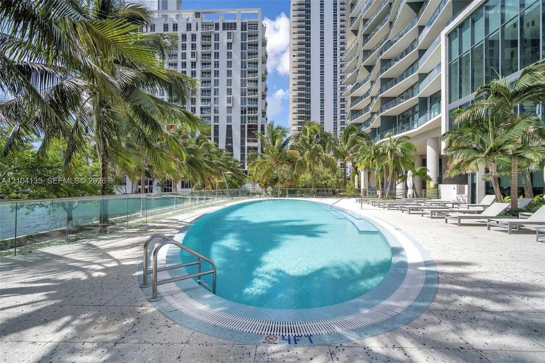 Oval-shaped swimming pool with a stainless steel ladder, surrounded by light-toned stone decking, glass panel perimeter fencing, and mature palm trees