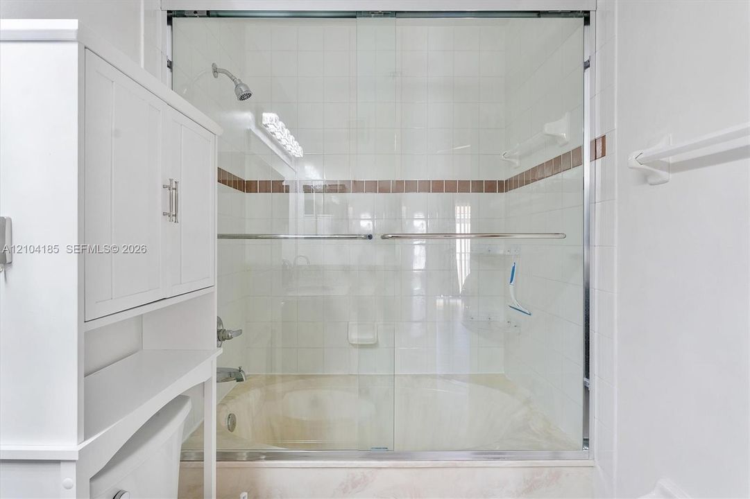 Shower and tub combination featuring a sliding glass enclosure, white ceramic tile surround with a decorative horizontal inlay, built-in shelving, and white cabinetry