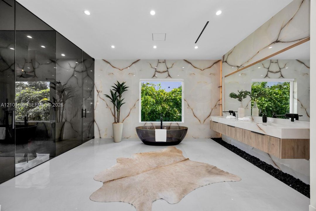 Full-wall stone slab surrounds, floating wood-finish dual vanity with integrated countertop, dark soaking tub, recessed ceiling lighting, and polished stone flooring
