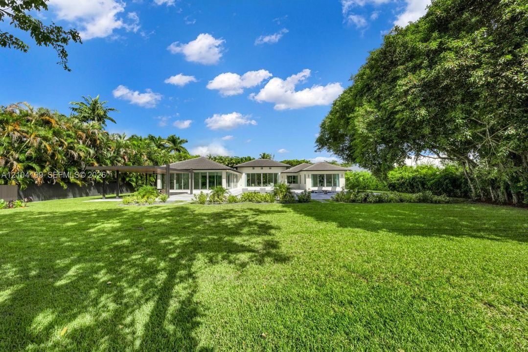Expansive backyard featuring a manicured lawn, perimeter landscaping, mature trees, a paved patio, and a pergola