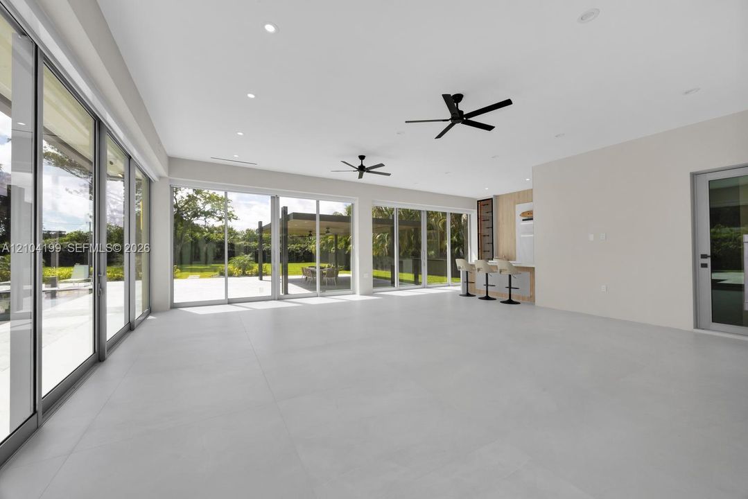 Large-format porcelain tile flooring, expansive floor-to-ceiling sliding glass doors, built-in wet bar with wood-panel detailing, recessed lighting, and dual matte black ceiling fans